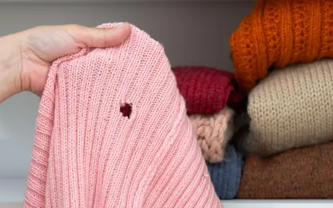Różowy sweter z dziurą, odkryty podczas przeglądania stosu ubrań w szafie. Widać inne swetry w różnych kolorach.