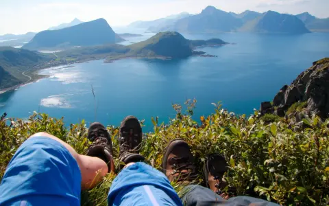 Fötter i vandringskängor vilar på en gräsbevuxen bergssluttning med en fantastisk utsikt över Lofoten. En perfekt dag för lofoten vandring.