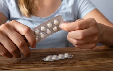 Dłonie trzymają opakowanie z tabletkami, które mogą być lekami z serotoniną. Na drewnianym blacie leży drugie, częściowo zużyte opakowanie.
