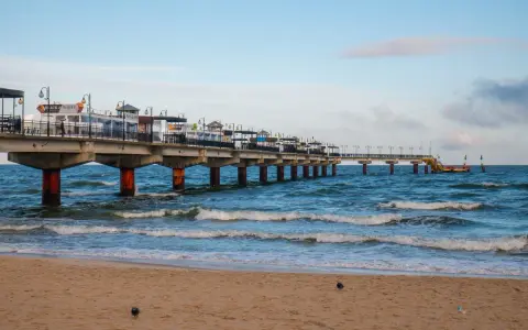 Molo w Międzyzdrojach, jedna z głównych atrakcji, wznosi się nad wzburzonym morzem. Piasek plaży i błękitne niebo tworzą malowniczy krajobraz.