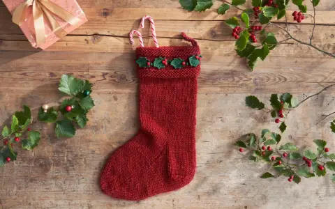 Röd stickad julstrumpa med grön misteldekoration och polkagrisar. En present och julgranspynt ligger på träbordet.