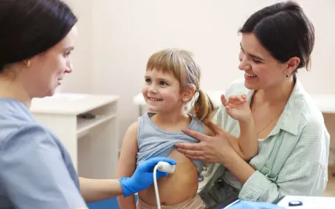Lekarz wykonuje usg brzuszka niemowlaka. Uśmiechnięta dziewczynka i jej mama towarzyszą badaniu.