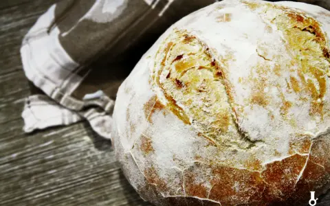 Chleb na suchych drożdżach, posypany mąką, z chrupiącą skórką, leży obok lnianej ściereczki na drewnianym blacie.