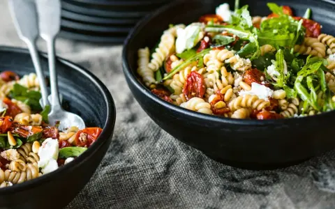 Zwei Schalen mit Nudelsalat mit getrockneten Tomaten, Pinienkernen und Rucola. Ein Löffel liegt in der vorderen Schale.