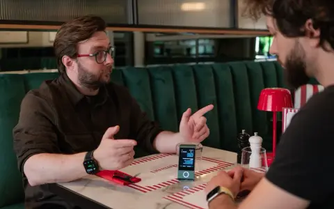 Robert Gryn, w okularach, gestykuluje podczas rozmowy przy stoliku w restauracji. Na stole stoi małe urządzenie z ekranem.