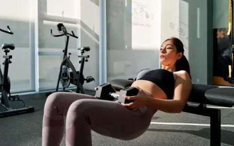 Femme enceinte effectuant un hip thrust au sol avec haltères, concentrée sur son entraînement dans une salle de sport lumineuse.