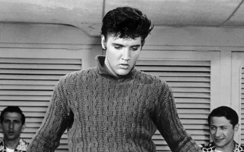 Elvis Presley in einem Strickpullover, vielleicht bei der Aufnahme seines letzten Liedes. Im Hintergrund zwei Männer.