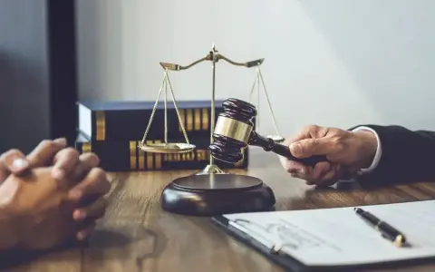 Dłoń trzymająca młotek sędziowski, waga sprawiedliwości i akta na drewnianym stole.
