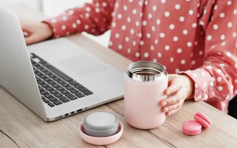 Kobieta przy laptopie trzyma kubek termiczny, obok leży nakrętka. Dowiedz się, jak wyczyścić nakrętkę od kubka termicznego, by napoje dłużej smakowały świeżo.