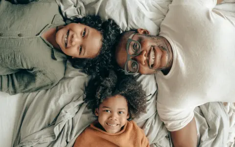 Un padre e i suoi due figli sorridono sdraiati sul letto, creando delle foto di famiglia originali e piene d'amore.