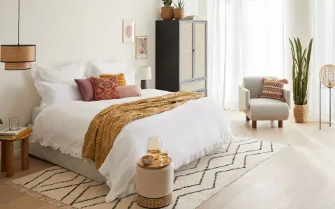 Habitación serena con cama blanca, manta ocre y cojines coloridos. Un armario de ratán y una silla acogedora complementan el estilo feng shui cama.