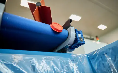Maszyna do produkcji folii, z niebieskim wałkiem i pomarańczową nakrętką, przygotowuje materiał cerplast do dalszej obróbki.