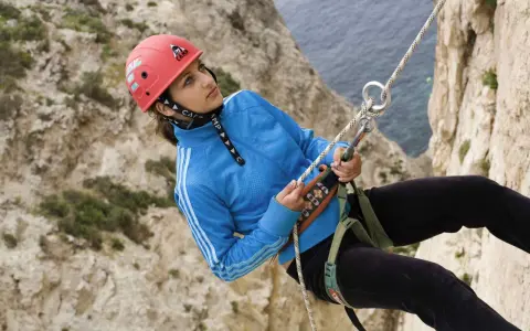 Junge Frau beim Abseilen an einer Felswand, Malta Klettern. Sie trägt einen roten Helm und eine blaue Jacke.