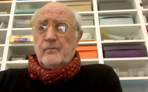 Ritratto di Francesco Pecoraro con occhiali gialli e sciarpa rossa, davanti a una libreria piena di scatole.