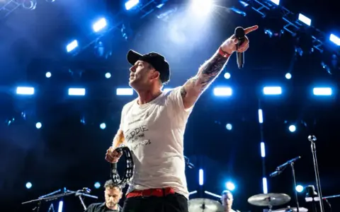 Wokalista z tatuażami i czapką na głowie prowadzi koncert One Republic, wskazując ręką w górę i trzymając tamburyn.