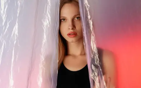 A young woman peeks through crinkled plastic sheet materials, her face illuminated by soft, colorful light.