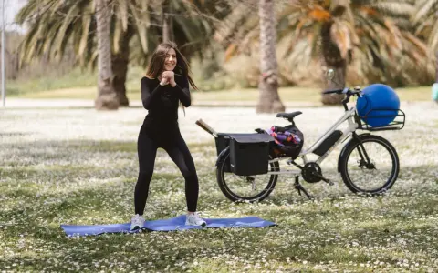 Donna sorridente fa esercizi all'aperto, accanto a una bicicletta per dimagrire con un grande pallone blu nel cestino.