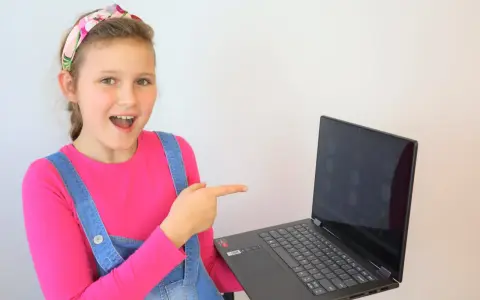 Uśmiechnięta dziewczynka w ogrodniczkach wskazuje na ekran laptopa, gdzie widać okienka z ikonami. Idealne miejsce na naukę angielskiego online dla dzieci.