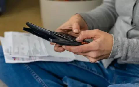 Ręce osoby w dżinsach i szarej bluzie liczą coś na kalkulatorze. Obok leżą dokumenty. Może to być rozliczenie rachunków, które zajmuje jej g12w godziny.