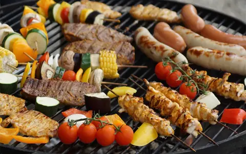 Una parrillada de que es bbq: brochetas de pollo y verduras, salchichas, bistecs y mazorcas de maíz asándose en una parrilla.