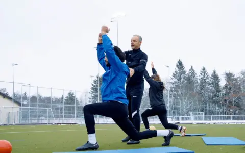 Athletik Training Fußball: Trainer korrigiert Ausfallschritt eines Spielers auf Kunstrasen. Zwei weitere Spieler machen Übungen im Hintergrund.