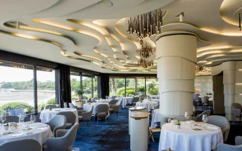 Salle élégante d'un restaurant chef de baie, tables dressées, vue sur la mer et jardin luxuriant. Plafond ondulé et luminaires modernes.