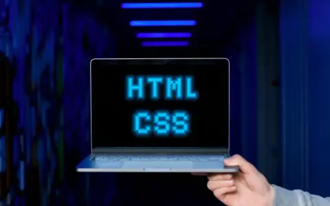 <p>Dłoń trzyma laptopa, na ekranie którego świeci napis "HTML CSS" w niebieskich pikselach. Tło jest ciemne, z niebieskimi neonami.</p>