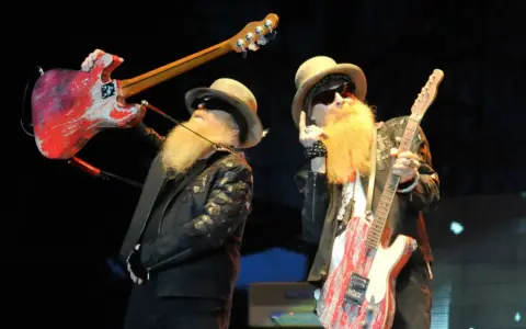 Dwóch muzyków z długimi, rudymi brodami, w kapeluszach i okularach, trzymających gitary. Jeden podnosi swoją gitarę nad głową, drugi pokazuje ją publiczności, tworząc wrażenie "over the under".