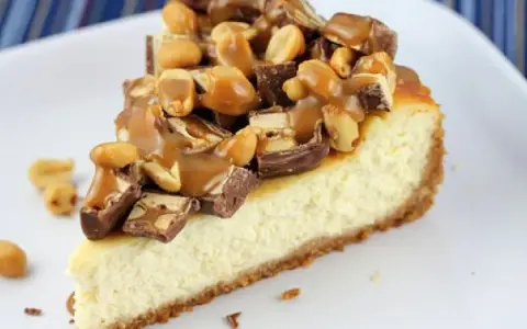 Kawałek sernika snickers z polewą karmelową i orzeszkami ziemnymi.