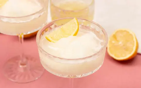 Erfrischendes Limoncello Sorbet in einem Kristallglas, garniert mit einer Zitronenspalte. Ein halbe Zitrone liegt daneben.