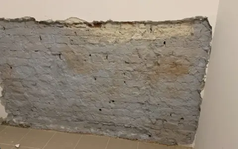 Fragment odsłoniętej, starej ceglanej ściany z szarym tynkiem i fragmentami żółtego podkładu.