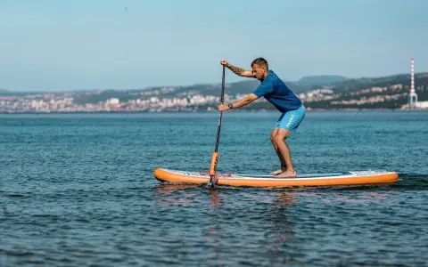 Mężczyzna na desce sup wiosłuje po spokojnym morzu, w tle widać miasto.