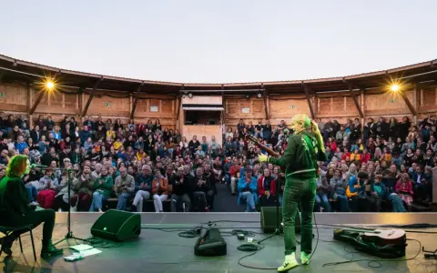 Konzert auf dem Tempelhofer Feld: Eine Musikerin spielt Gitarre vor einem begeisterten Publikum in einem hölzernen Amphitheater. Ein wahrer luftschloss tempelhofer feld Traum.