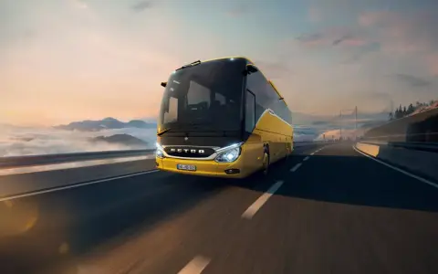 Autobús Setra amarillo brillante viaja por una carretera escénica al amanecer, con nubes y montañas de fondo.