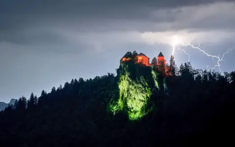 Zamek Poenari, oświetlony na czerwono, góruje nad skalistym zboczem podczas burzy z piorunami.