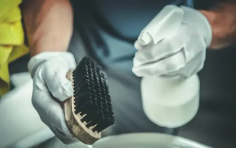 Manos enguantadas sostienen cepillo y spray. Preparación para la limpieza de estufa de pellets, consulta el precio limpieza estufa pellets.