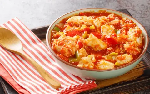 Un tazón de bacalao ajoarriero con pimientos y tomate, servido con una cuchara de madera sobre una servilleta a rayas.