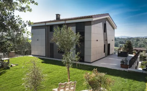 Esterno di case moderne con facciata in pannelli chiari e scuri, giardino curato e vista panoramica.