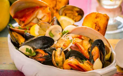 Un fumetto di pesce ricco di cozze, vongole e crostacei, guarnito con limone e prezzemolo, servito in una ciotola bianca.