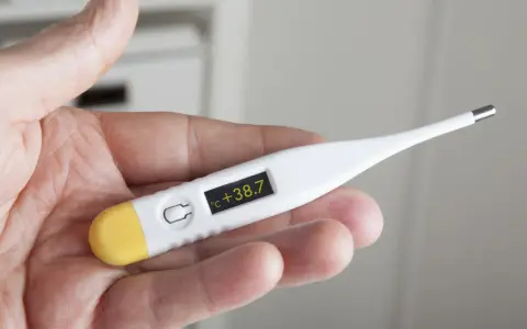 Dłoń trzyma termometr wskazujący 38.7°C. Czas na działanie, jak zbić gorączkę.
