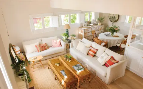 Salón luminoso con sofás blancos, mesas de mimbre y un rincón de comedor. La distribución de salones irregulares crea un espacio acogedor y funcional.
