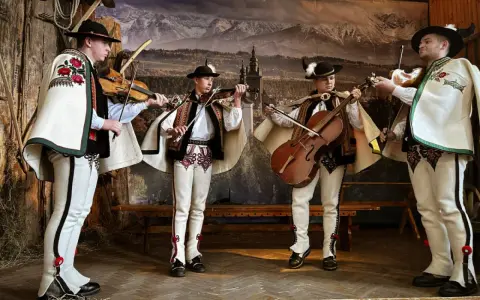 Czterech muzykantów w strojach góralskich gra na skrzypcach i basach. Gęśliki podhalańskie brzmią w tle górskiego krajobrazu.