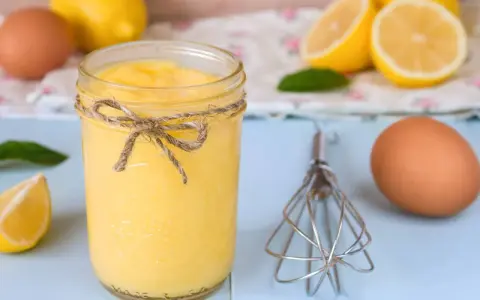 Un tarro de lemon curd casero, atado con cuerda, junto a limones, huevos y un batidor.