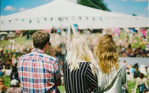 Tre vänner njuter av en solig dag på en musikfestival i Stockholm. En stor vit tältduk och flaggor syns i bakgrunden.
