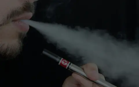 Un hombre con barba exhala vapor de un vapeador, similar al estilo Heisenberg.