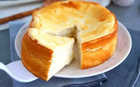 Un délicieux gâteau fromage blanc, doré et moelleux, servi sur une assiette blanche. Une part est déjà coupée.