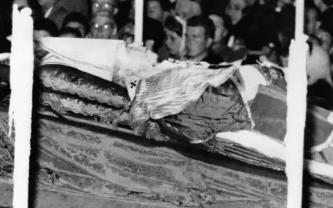 Pius XII w szatach, przygotowany do balsamowania. Tłum ludzi w tle.