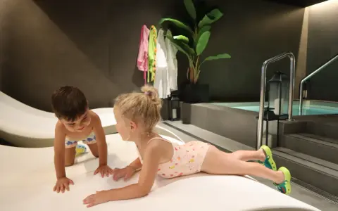 Niños jugando en un spa para niños, con una piscina al fondo y toallas colgadas.