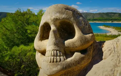 Escultura de calavera en la **ruta de las caras**, con un lago azul y colinas verdes al fondo.