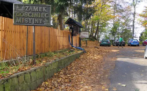 Tablica z napisem "Zamek GOSCINIEC MISYJNY" przy drewnianym płocie. Jesienny parking z samochodami i opadłymi liśćmi.